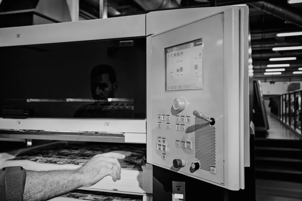Black-and-white detail of a printing press control panel with an operator handling a printed sheet
