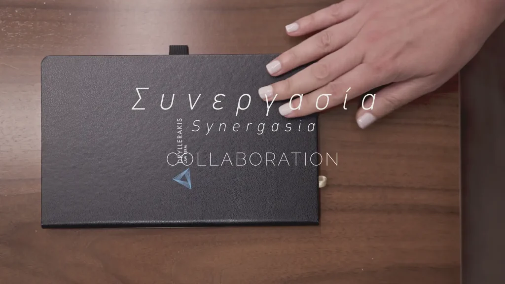 Hand resting on a black notebook with Συνεργασία and Collaboration overlaid in the ROOTS brand film