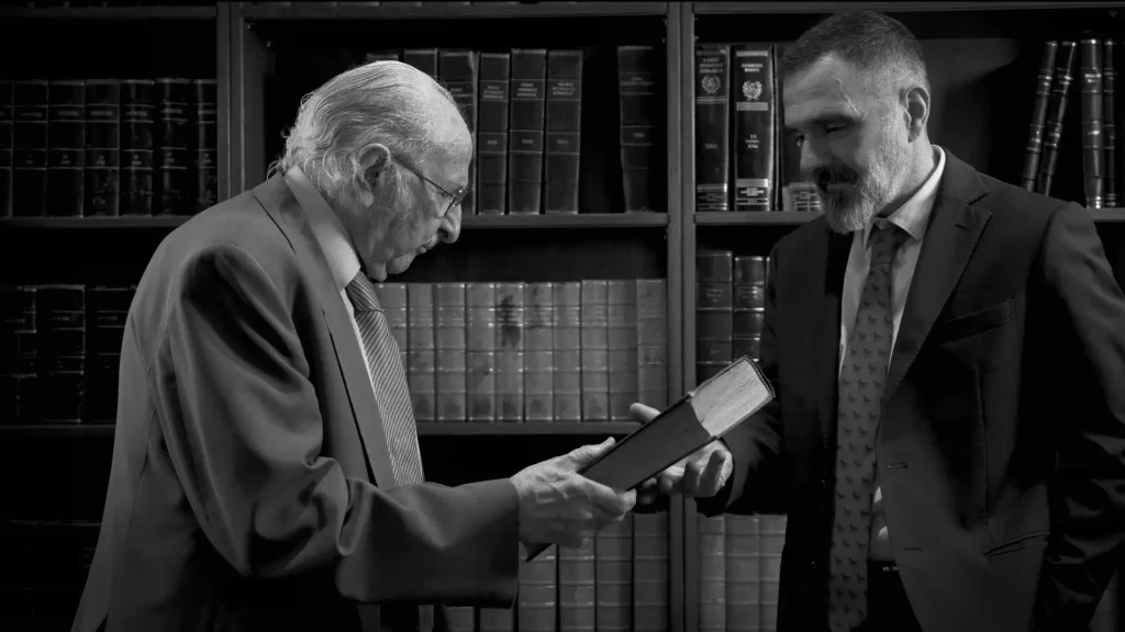 Black and white library scene with senior Dryllerakis handing a book to Emmanuel Dryllerakis as an inheritance gesture